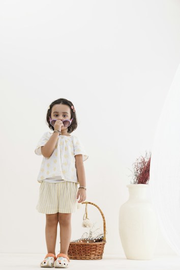A young child wearing a light-colored outfit stands indoors, holding sunglasses to their face. Beside them is a wicker basket filled with dried plants, and a large white vase also containing dried stems. The setting is minimalist with neutral colors.