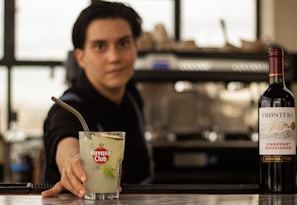 A bartender offering a cocktail in a glass with a Havana Club logo, accompanied by a bottle of Frontera Cabernet Sauvignon on the counter, with a blurred background.