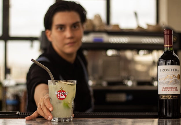 A bartender offering a cocktail in a glass with a Havana Club logo, accompanied by a bottle of Frontera Cabernet Sauvignon on the counter, with a blurred background.