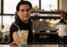 A bartender offering a cocktail in a glass with a Havana Club logo, accompanied by a bottle of Frontera Cabernet Sauvignon on the counter, with a blurred background.