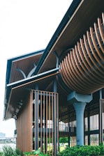 A modern architectural structure with a unique and intricate design featuring wooden slats and metal elements. The building incorporates both vertical and curved components, creating a striking visual contrast. Lush greenery surrounds the base, adding a natural element to the contemporary design.