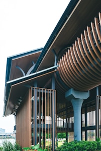 A modern architectural structure with a unique and intricate design featuring wooden slats and metal elements. The building incorporates both vertical and curved components, creating a striking visual contrast. Lush greenery surrounds the base, adding a natural element to the contemporary design.