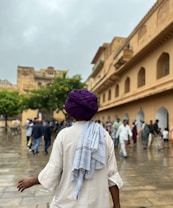 A person wearing a purple turban and light-colored traditional attire is seen from behind, walking along a wet stone pathway. In the background, a historic building with arches and a crowd of people are visible, along with trees under a cloudy sky.