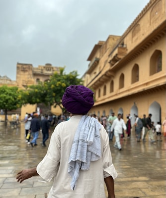 A person wearing a purple turban and light-colored traditional attire is seen from behind, walking along a wet stone pathway. In the background, a historic building with arches and a crowd of people are visible, along with trees under a cloudy sky.
