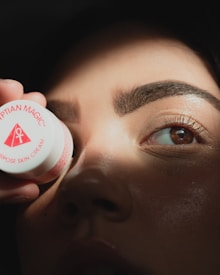 A close-up of a person's face partially illuminated by light, with a focus on the eyes and nose. They are holding a small container labeled 'Egyptian Magic' near the eye.