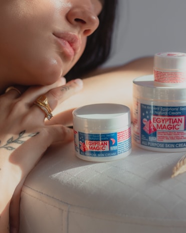 A person with light skin tone, wearing a gold ring, leans on a surface next to two jars of Egyptian Magic all-purpose skin cream. The focus is on the cream and the person's hand, which features a tattoo with delicate linework.