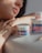 A person with light skin tone, wearing a gold ring, leans on a surface next to two jars of Egyptian Magic all-purpose skin cream. The focus is on the cream and the person's hand, which features a tattoo with delicate linework.