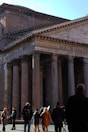 A group of people are gathered in front of an ancient Roman building, characterized by its grand columns and large stone facade. The structure includes the inscription 'M. AGRIPPA L. F.' on the pediment. The scene is bustling with activity as tourists explore the area.