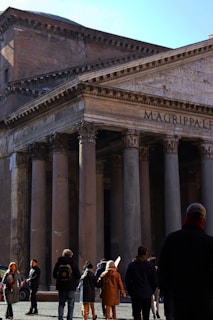 A group of people are gathered in front of an ancient Roman building, characterized by its grand columns and large stone facade. The structure includes the inscription 'M. AGRIPPA L. F.' on the pediment. The scene is bustling with activity as tourists explore the area.