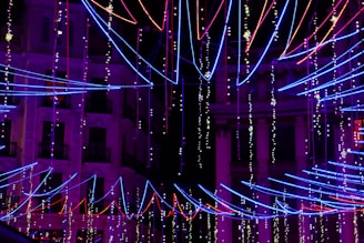 Colorful neon lights in red, blue, and purple form intricate patterns against the backdrop of a darkened building. The strings of lights are arranged in diagonal lines and other geometric formations, creating a vibrant and festive atmosphere.