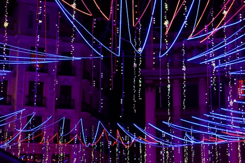 Colorful neon lights in red, blue, and purple form intricate patterns against the backdrop of a darkened building. The strings of lights are arranged in diagonal lines and other geometric formations, creating a vibrant and festive atmosphere.