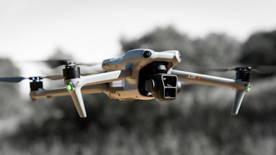 A quadcopter drone in flight, featuring four propellers attached to a sleek, metallic body. The camera at the front is prominently visible, and the background is slightly blurred with natural elements, suggesting it is outdoors.