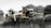 A quadcopter drone in flight, featuring four propellers attached to a sleek, metallic body. The camera at the front is prominently visible, and the background is slightly blurred with natural elements, suggesting it is outdoors.