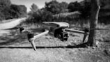 A drone is hovering in mid-air against a background of blurred outdoor scenery with trees and a dirt path. The image is in black and white, emphasizing the sleek design of the drone with its propellers visible.