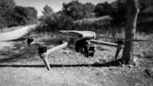 A drone is hovering in mid-air against a background of blurred outdoor scenery with trees and a dirt path. The image is in black and white, emphasizing the sleek design of the drone with its propellers visible.
