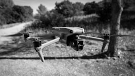 A drone is hovering in mid-air against a background of blurred outdoor scenery with trees and a dirt path. The image is in black and white, emphasizing the sleek design of the drone with its propellers visible.
