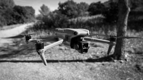 A drone is hovering in mid-air against a background of blurred outdoor scenery with trees and a dirt path. The image is in black and white, emphasizing the sleek design of the drone with its propellers visible.