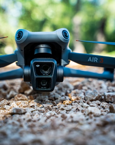 A close-up view of a drone resting on a rocky surface with a blurred green natural background. The focus is on the drone's camera system and front arms, featuring prominent branding and technical specifications.