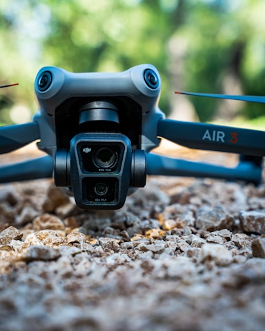 A close-up view of a drone resting on a rocky surface with a blurred green natural background. The focus is on the drone's camera system and front arms, featuring prominent branding and technical specifications.