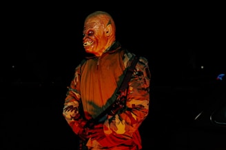 A person wearing a monstrous mask stands in the dark, illuminated by an eerie red light. The figure is dressed in a camouflaged jacket and holds a strap across their body, creating a menacing appearance.