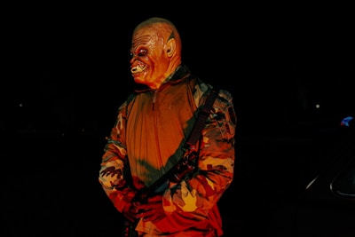 A person wearing a monstrous mask stands in the dark, illuminated by an eerie red light. The figure is dressed in a camouflaged jacket and holds a strap across their body, creating a menacing appearance.