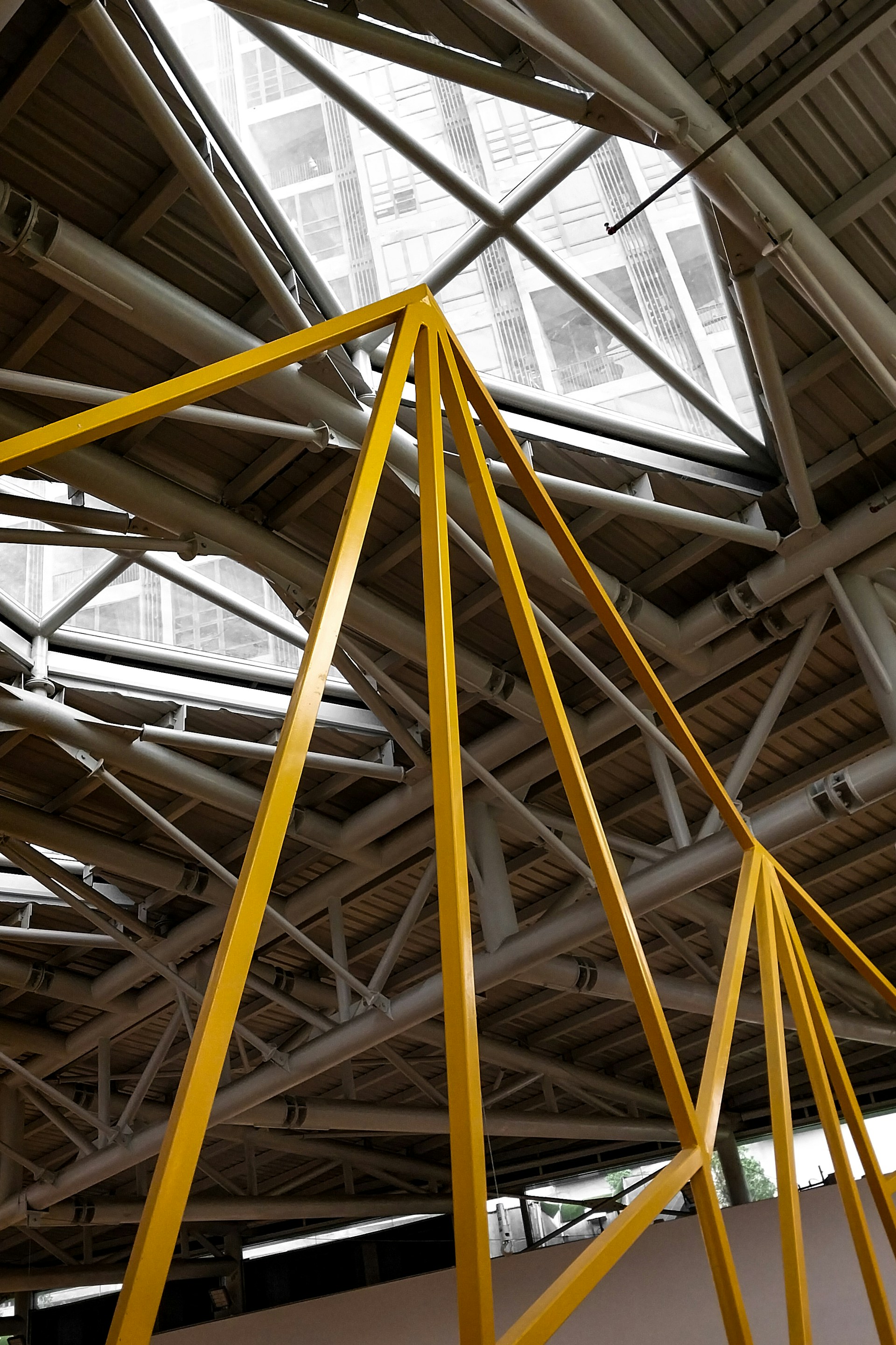 A modern architectural structure featuring a geometric metal framework with yellow beams set against a background of exposed ceiling pipes and a glass roof. The ceiling is composed of various intersecting metal beams creating a complex pattern, and natural light filters through the glass overhead.