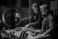 Two DJs are performing in a dimly lit environment with a disco ball reflecting light around the room. They are focused on their equipment, wearing headphones, and surrounded by plants and DJ gear.