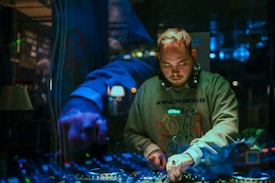 A person with headphones is focused on mixing music at a DJ booth. The setting is dimly lit, with reflections adding a dynamic, layered effect. The background includes colorful lights and equipment, creating an engaging nightlife atmosphere.