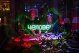 Two DJs are performing in a dimly lit club environment, surrounded by green and red lighting. One DJ is wearing headphones while operating the DJ equipment. A digital display shows text in a vibrant green above the DJ booth. The setup is decorated with plants, adding to the lively atmosphere.