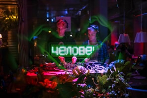 Two DJs are performing in a dimly lit club environment, surrounded by green and red lighting. One DJ is wearing headphones while operating the DJ equipment. A digital display shows text in a vibrant green above the DJ booth. The setup is decorated with plants, adding to the lively atmosphere.
