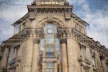 A historic architectural building with ornate details and large columns is reflected in a window, creating a layered visual effect. The building features classical design elements, including decorative stone carvings and arched windows. Above the main entrance is a clock and a sign in Cyrillic script.