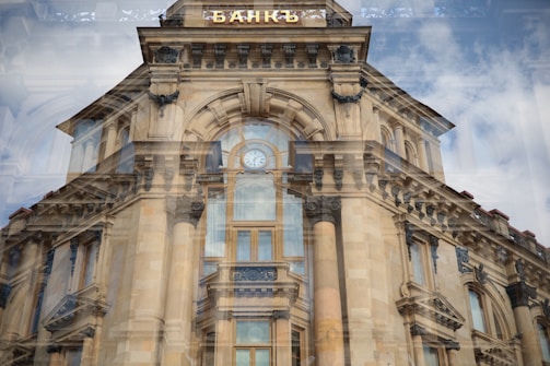 A historic architectural building with ornate details and large columns is reflected in a window, creating a layered visual effect. The building features classical design elements, including decorative stone carvings and arched windows. Above the main entrance is a clock and a sign in Cyrillic script.