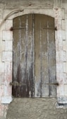 A rustic wooden shutter closed over a window set within a stone wall. The wood appears weathered and worn, with vertical panels showing peeling paint and aging marks. Surrounding the shutter is a white stone frame, also showing signs of decay with a rough, textured surface. A metal pipe curves above the shutter, adding to the rustic and aged aesthetic.