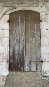 A rustic wooden shutter closed over a window set within a stone wall. The wood appears weathered and worn, with vertical panels showing peeling paint and aging marks. Surrounding the shutter is a white stone frame, also showing signs of decay with a rough, textured surface. A metal pipe curves above the shutter, adding to the rustic and aged aesthetic.