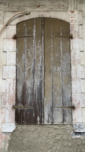 A rustic wooden shutter closed over a window set within a stone wall. The wood appears weathered and worn, with vertical panels showing peeling paint and aging marks. Surrounding the shutter is a white stone frame, also showing signs of decay with a rough, textured surface. A metal pipe curves above the shutter, adding to the rustic and aged aesthetic.