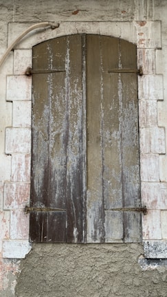 A rustic wooden shutter closed over a window set within a stone wall. The wood appears weathered and worn, with vertical panels showing peeling paint and aging marks. Surrounding the shutter is a white stone frame, also showing signs of decay with a rough, textured surface. A metal pipe curves above the shutter, adding to the rustic and aged aesthetic.