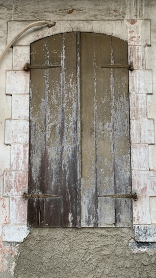 A rustic wooden shutter closed over a window set within a stone wall. The wood appears weathered and worn, with vertical panels showing peeling paint and aging marks. Surrounding the shutter is a white stone frame, also showing signs of decay with a rough, textured surface. A metal pipe curves above the shutter, adding to the rustic and aged aesthetic.