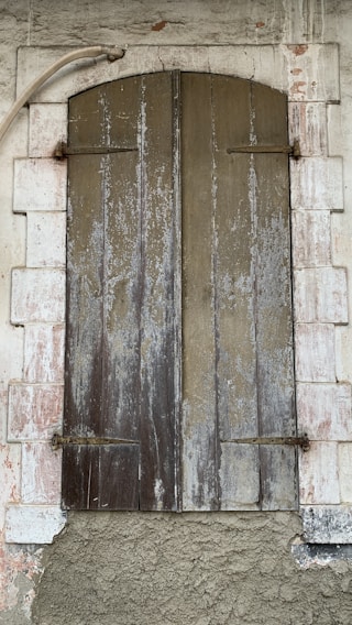 A rustic wooden shutter closed over a window set within a stone wall. The wood appears weathered and worn, with vertical panels showing peeling paint and aging marks. Surrounding the shutter is a white stone frame, also showing signs of decay with a rough, textured surface. A metal pipe curves above the shutter, adding to the rustic and aged aesthetic.