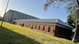 A perspective view of two adjacent buildings. The foreground features a single-story brick structure with a dark roof and several evenly spaced windows. Behind it, a taller multi-story building with numerous windows extends into the sky. The setting is a grassy area with trees and a street lamp, suggesting an urban or institutional environment. Sunlight casts shadows, indicating an early morning or late afternoon.