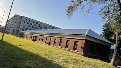 A perspective view of two adjacent buildings. The foreground features a single-story brick structure with a dark roof and several evenly spaced windows. Behind it, a taller multi-story building with numerous windows extends into the sky. The setting is a grassy area with trees and a street lamp, suggesting an urban or institutional environment. Sunlight casts shadows, indicating an early morning or late afternoon.