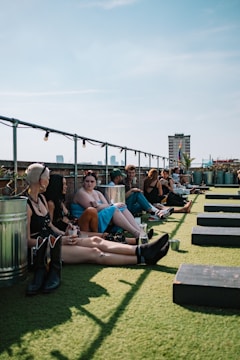 A group of people sitting and lounging on a rooftop with artificial grass. They are casually dressed and appear to be socializing. There are potted plants and city buildings visible in the background. The weather is clear and sunny, casting shadows on the grass.