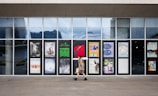 A person walks past a series of glass panels with various posters and advertisements displayed on them. The background shows a modern urban environment reflected in the glass.