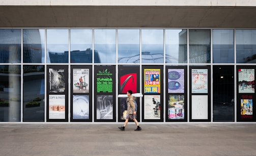 A person walks past a series of glass panels with various posters and advertisements displayed on them. The background shows a modern urban environment reflected in the glass.