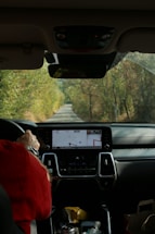 The interior of a car showing the dashboard with a navigation system displaying a map. A person wearing a red jacket is seated in the passenger seat, holding a soda can, with foliage visible through the windshield lining the narrow road ahead.