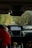 The interior of a car showing the dashboard with a navigation system displaying a map. A person wearing a red jacket is seated in the passenger seat, holding a soda can, with foliage visible through the windshield lining the narrow road ahead.