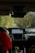 The interior of a car showing the dashboard with a navigation system displaying a map. A person wearing a red jacket is seated in the passenger seat, holding a soda can, with foliage visible through the windshield lining the narrow road ahead.