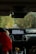 The interior of a car showing the dashboard with a navigation system displaying a map. A person wearing a red jacket is seated in the passenger seat, holding a soda can, with foliage visible through the windshield lining the narrow road ahead.