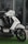 A modern, sleek-looking scooter features a white body with black accents, positioned on a patch of artificial green grass. The scooter has a futuristic design with its headlight prominent and handlebars visible. The background includes a reflective glass window, hinting at a modern urban setting.