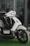 A modern, sleek-looking scooter features a white body with black accents, positioned on a patch of artificial green grass. The scooter has a futuristic design with its headlight prominent and handlebars visible. The background includes a reflective glass window, hinting at a modern urban setting.