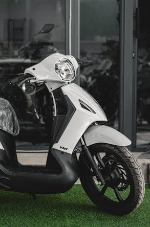 A modern, sleek-looking scooter features a white body with black accents, positioned on a patch of artificial green grass. The scooter has a futuristic design with its headlight prominent and handlebars visible. The background includes a reflective glass window, hinting at a modern urban setting.